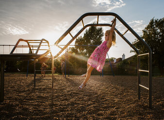 Side view of girl hanging on monkey bars against sky at playground during sunset - CAVF51514