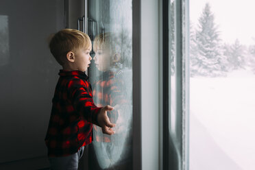 Seitenansicht eines Jungen, der durch das Fenster schaut, während er im Winter zu Hause steht - CAVF51539
