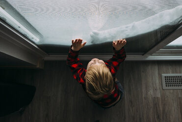 Hoher Blickwinkel des Jungen, der durch das Fenster schaut, während er zu Hause im Winter steht - CAVF51540