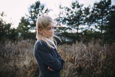 Side view of thoughtful girl with arms crossed standing on grassy field - CAVF51541