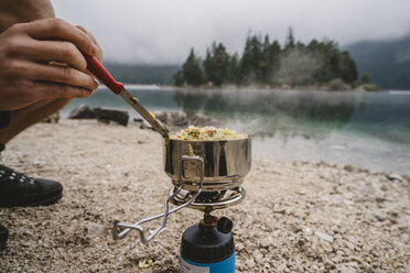 Tiefschnitt eines Mannes, der am Seeufer im Wald kauernd Essen auf einem Campingkocher zubereitet - CAVF51567