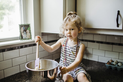 Porträt eines niedlichen Mädchens, das Essen in einem Behälter zubereitet, während es auf der Küchentheke zu Hause sitzt, lizenzfreies Stockfoto
