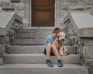 Junge umarmt Hund, während er auf einer Treppe sitzt - CAVF51624