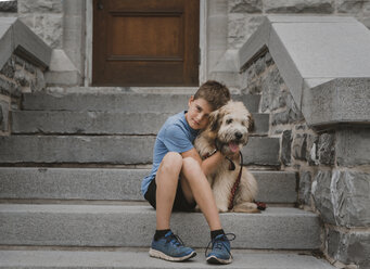 Porträt eines Jungen, der auf einer Treppe sitzend einen Hund umarmt - CAVF51625
