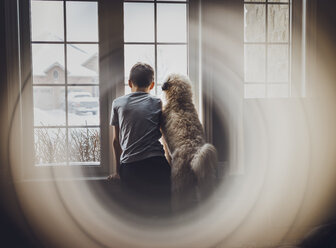 Rückansicht eines Jungen mit Hund, der durch ein Fenster schaut, gesehen durch ein Glas zu Hause - CAVF51626