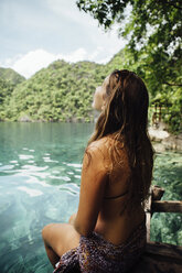 Carefree young woman sitting by sea against mountains - CAVF51636