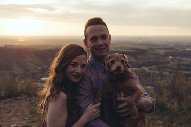 Porträt eines jungen Paares mit Hund, das bei Sonnenuntergang auf einem Feld steht - CAVF51668