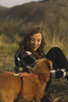 Lächelnde junge Frau sieht ihren Hund an, während sie auf einem Feld sitzt - CAVF51681