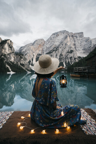 Seitenansicht einer jungen Frau mit beleuchteter Lichterkette und Laterne, die am See vor den Bergen sitzt, lizenzfreies Stockfoto