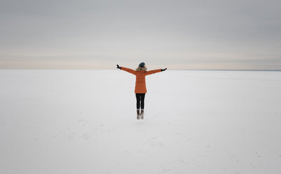 Rückansicht einer Frau mit ausgestreckten Armen, die auf einer verschneiten Landschaft vor einem bewölkten Himmel steht - CAVF51721