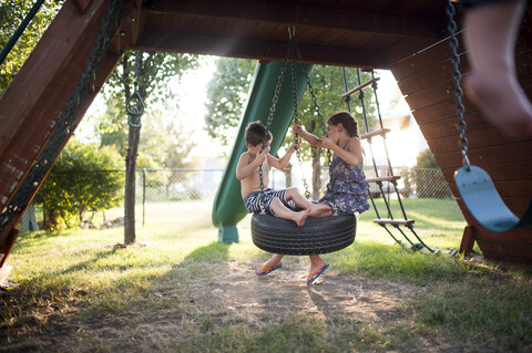 Verspielte Geschwister schwingen auf einer Reifenschaukel auf dem Spielplatz, lizenzfreies Stockfoto