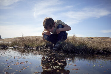 Boy looking into lake while crouching on grassy field against sky - CAVF51912
