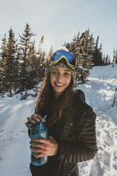 Porträt einer lächelnden jungen Frau, die eine Skibrille trägt und eine Wasserflasche auf einem verschneiten Feld hält - CAVF51935