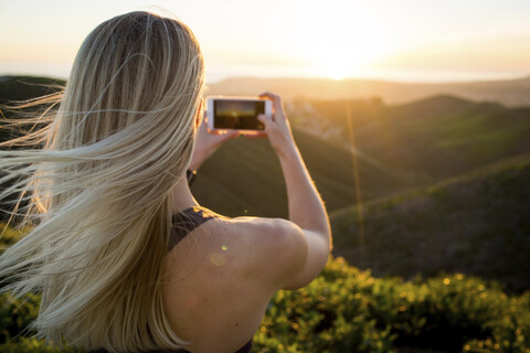 Rückansicht einer Frau, die während des Sonnenuntergangs auf einem Berg steht und mit ihrem Handy fotografiert, lizenzfreies Stockfoto