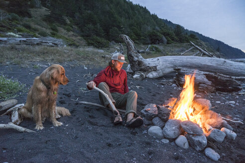 Mann und Hund sitzen am Lagerfeuer am Strand - CAVF51957