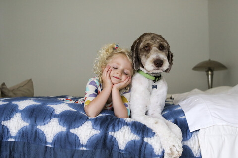 Mädchen mit geschlossenen Augen und Händen am Kinn liegt neben einem Hund auf einem Bett an der Wand, lizenzfreies Stockfoto