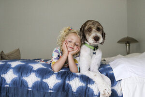 Mädchen mit geschlossenen Augen und Händen am Kinn liegt neben einem Hund auf einem Bett an der Wand - CAVF51967