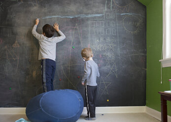 Rear view of brothers drawing with chalk on blackboard wall at home - CAVF52005