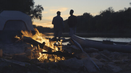 Silhouette von Vater und Sohn am Seeufer stehend mit brennendem Lagerfeuer im Vordergrund - CAVF52018