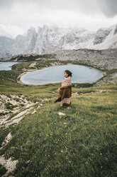 Side view of woman with blanket standing on grassy field by lake against cloudy sky - CAVF52045