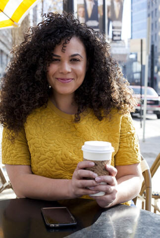 Porträt einer lächelnden Frau, die einen Einwegbecher hält, während sie in einem Straßencafé in der Stadt sitzt, lizenzfreies Stockfoto