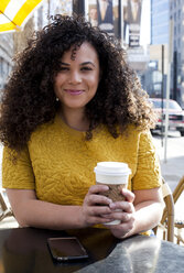 Portrait of smiling woman holding disposable cup while sitting at sidewalk cafe in city - CAVF52078