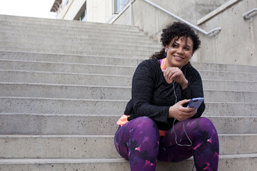 Niedriger Blickwinkel einer Frau, die auf einer Treppe sitzend mit ihrem Handy Musik hört - CAVF52079