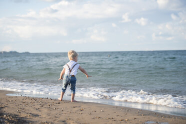 Rückansicht eines kleinen Jungen, der einen Stein ins Meer wirft, während er am Strand vor einem bewölkten Himmel steht - CAVF52098