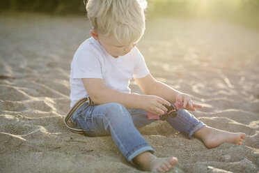 Ein kleiner Junge spielt mit einem Spielzeug, während er im Sand am Strand sitzt - CAVF52099