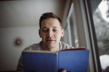 Niedriger Blickwinkel eines Mannes, der ein Buch liest, während er zu Hause am Fenster sitzt - CAVF52152