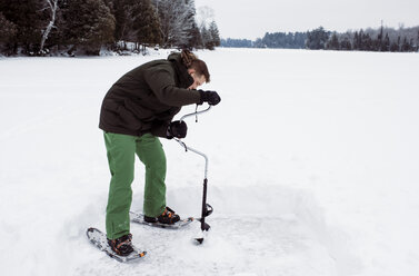 Man drilling frozen lake with ice auger standing against sky - CAVF52154