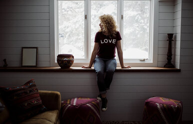 Frau in voller Länge mit LOVE-Text-T-Shirt beim Blick durch das Fenster zu Hause - CAVF52158