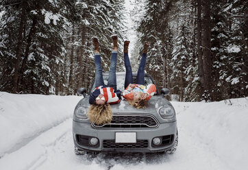 Verspielte Freundinnen in voller Länge auf der Motorhaube eines Autos liegend und mit hochgelegten Füßen im Winter - CAVF52161