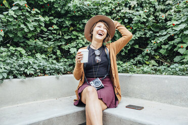 Cheerful woman holding disposable cup while sitting on bench at park - CAVF52221
