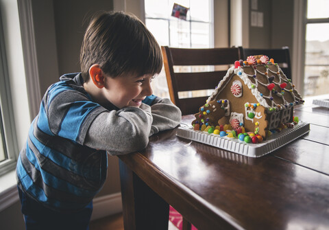 Glücklicher Junge, der ein Lebkuchenhaus auf einem Holztisch in der Weihnachtszeit betrachtet, lizenzfreies Stockfoto