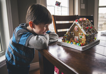 Glücklicher Junge, der ein Lebkuchenhaus auf einem Holztisch in der Weihnachtszeit betrachtet - CAVF52233