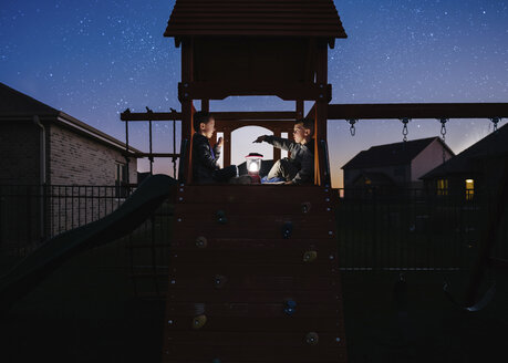 Brüder mit beleuchteter Laterne sitzen auf einem Spielgerät im Freien vor einem Sternenfeld im Park - CAVF52267