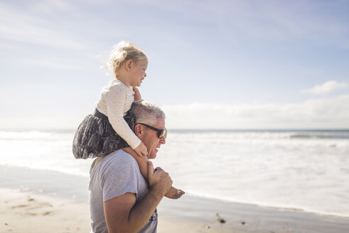 Seitenansicht des Großvaters, der seine Enkelin auf den Schultern trägt, während er am Strand vor dem Himmel steht - CAVF52268
