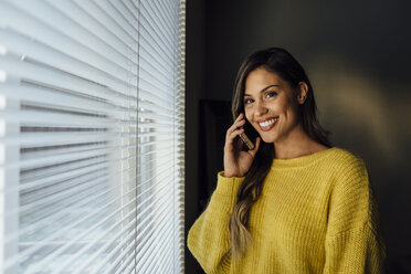 Porträt einer lächelnden Frau, die zu Hause am Fenster steht und mit ihrem Handy spricht - CAVF52273