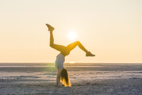 Seitenansicht der Frau macht Handstand am Strand gegen klaren Himmel bei Sonnenuntergang, lizenzfreies Stockfoto