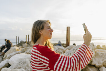 Seitenansicht einer jungen Frau, die ein Selfie mit ihrem Smartphone am Strand macht - CAVF52334