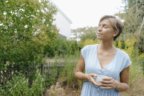 Frau mit geschlossenen Augen steht im Garten und hält eine Tasse Kaffee - KNSF05055