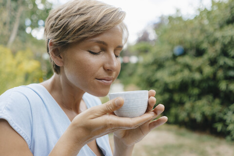 Porträt einer Frau mit geschlossenen Augen im Garten, die eine Tasse Kaffee hält, lizenzfreies Stockfoto