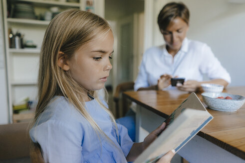 Mädchen mit Buch sitzt am Tisch zu Hause mit Mutter mit Handy im Hintergrund - KNSF05072
