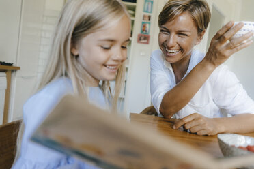Glückliche Mutter und Tochter mit Buch am Tisch sitzend zu Hause - KNSF05073