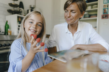 Happy mother and daughter sitting at table in kitchen at home playing with raspberries - KNSF05078