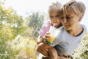 Mutter trägt Tochter huckepack im Garten und trinkt einen Smoothie - KNSF05097