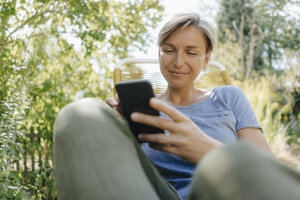 Woman sitting in garden on chair using cell phone - KNSF05111