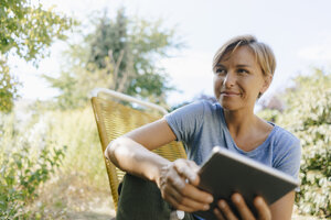 Woman sitting in garden on chair holding tablet - KNSF05125