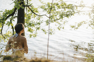 Rückansicht einer Frau im Bikini an einem Baumstamm an einem See - KNSF05172